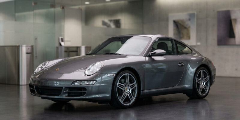 Porsche 911 gris anthracite (type 997) stationnée dans un showroom lumineux avec des murs vitrés et œuvres d'art.